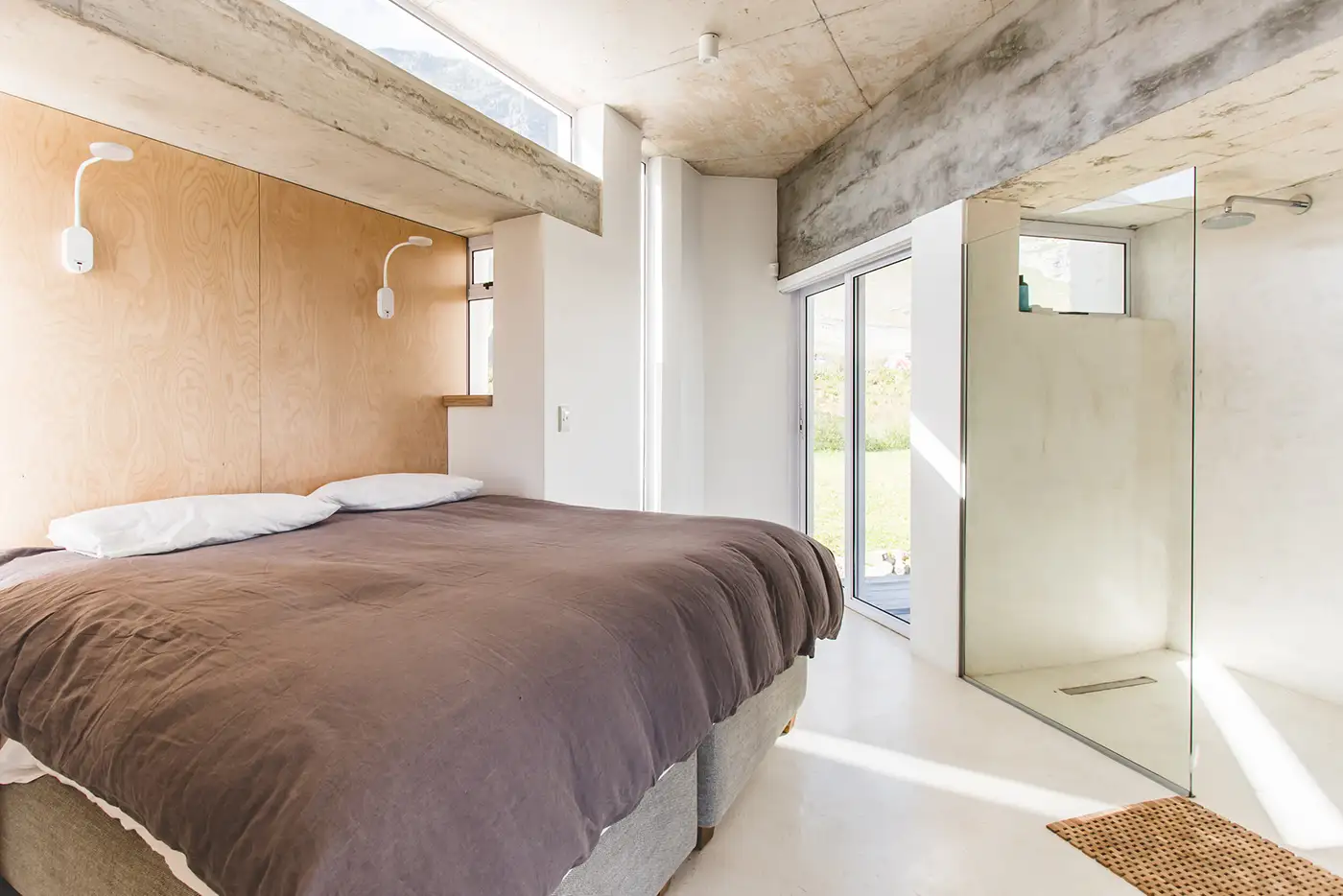 Bedroom with a concrete ceiling, plywood headboard wall and glass shower